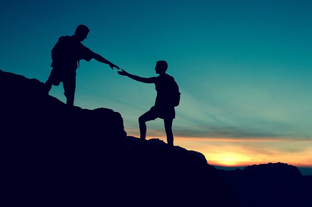 couple-climbing-help-5946815 Ein Mann auf einem Berg der seine Hand ausstreckt um einem anderen Mann zu helfen den Berg raufzuwandern.. Symbolik für Hilfe bei Spielsucht.